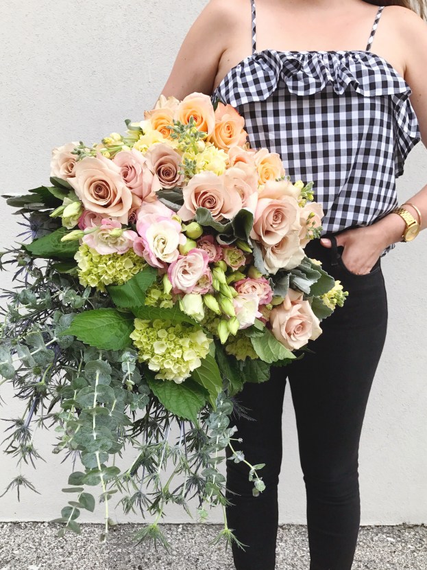 Florist in gingham camisole holding a large bundle of fresh flowers including roses, hydrangea, fresh eucalyptus, dusty miller, stock flower and blue thistle.