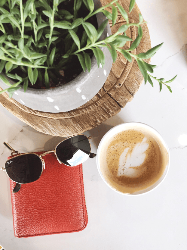 Coffee shop flat lay with Ray-Ban hexagonal sunglasses, Cuyana small zip around wallet, and cappuccino